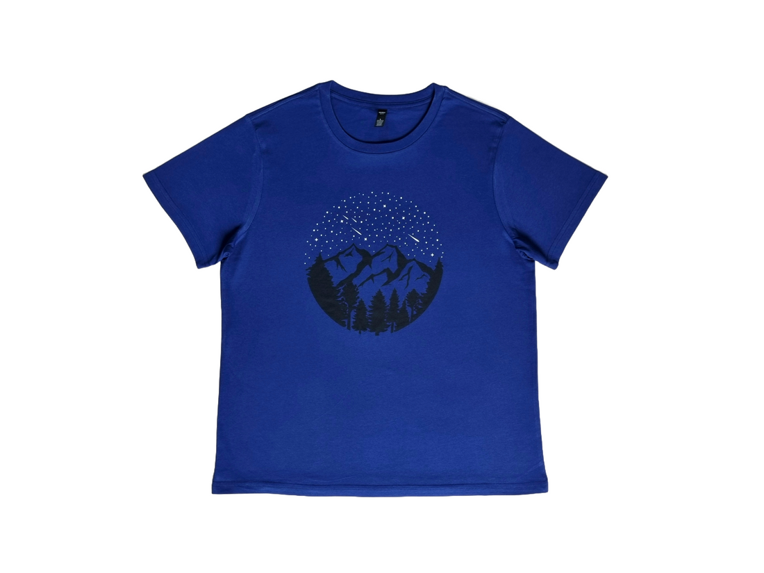 A dark blue women's tee shirt with a design featuring a starry sky and shooting stars, over mountains and trees, printed with eco friendly inks.